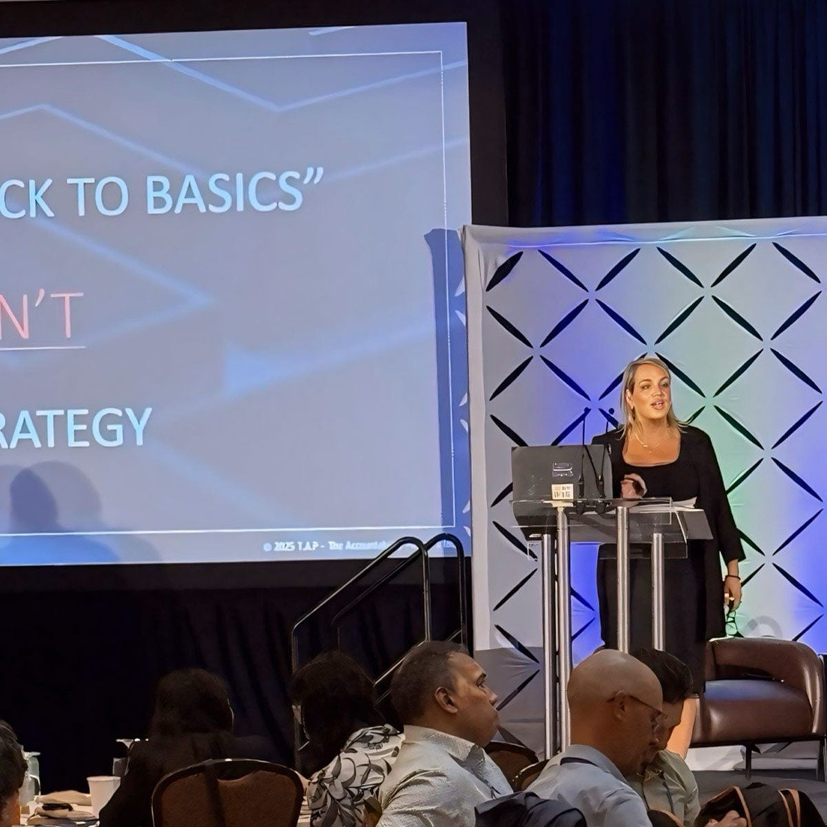 Woman giving a presentation at a conference with a screen displaying 'Back to Basics' strategy.