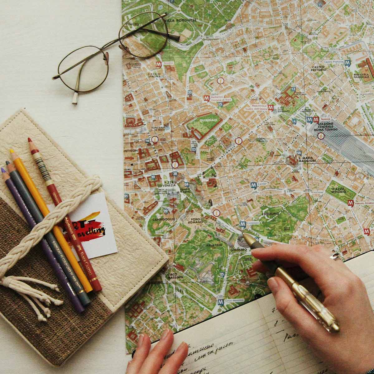 Person holding a pen over a city map with glasses and coloring books on a table.
