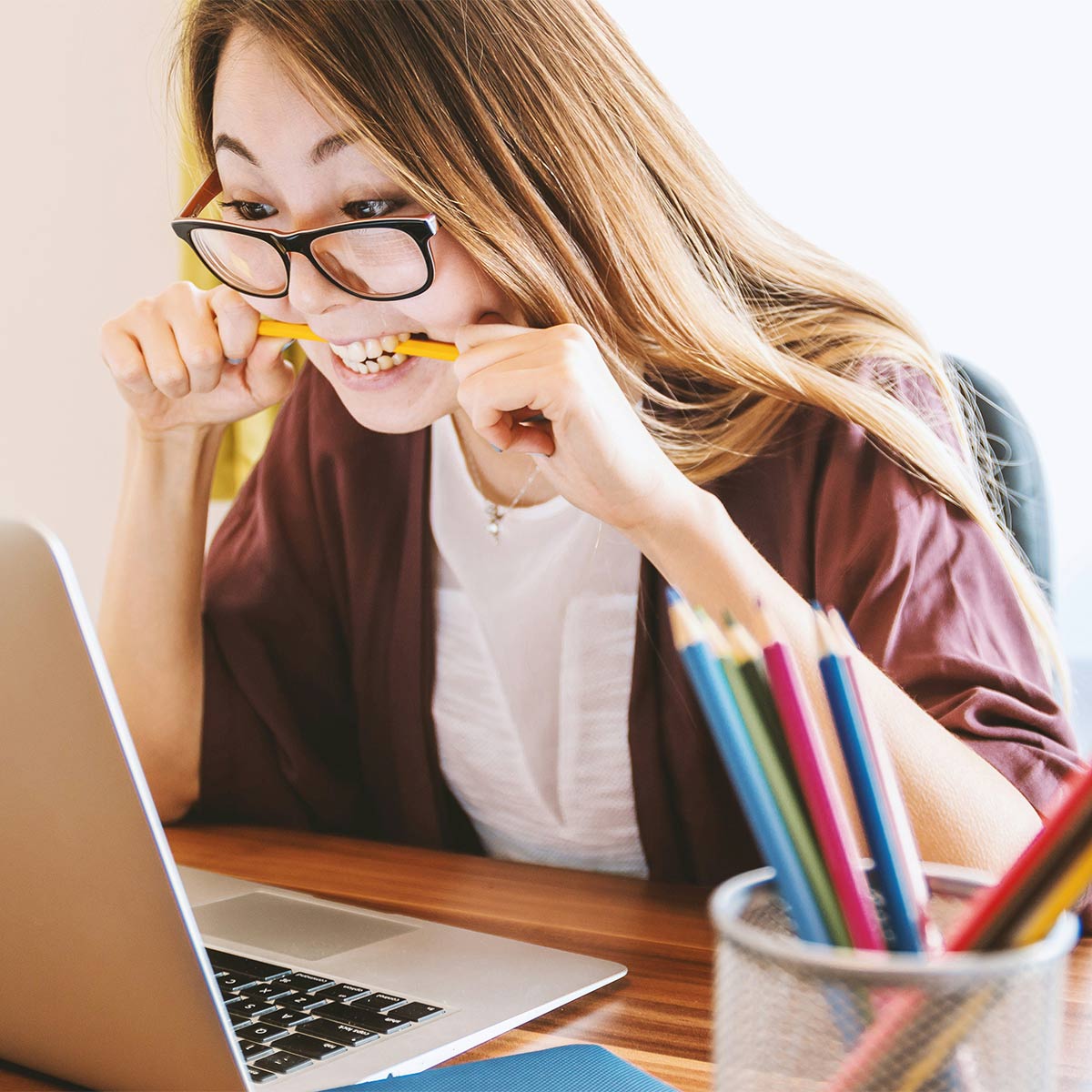 Person sitting at a desk with a laptop, holding a pencil to their mouth, with a container of colored pencils on the desk.