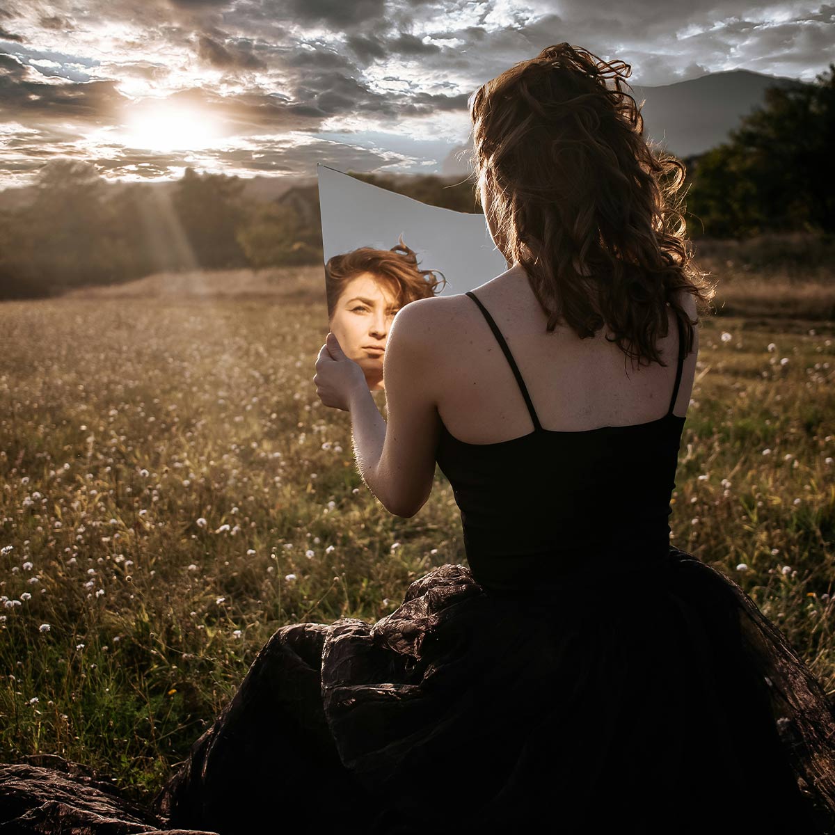 Woman in a black dress holding and looking into a mirror in a field with mountains and sunlit clouds.