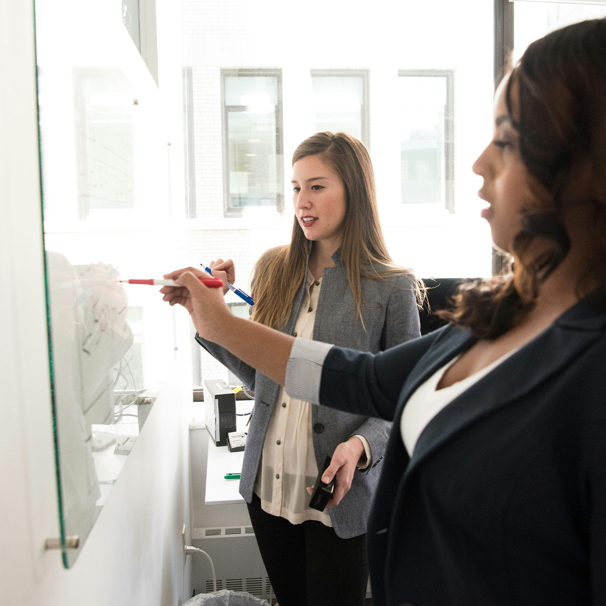 Two women in a professional setting, one writing on a whiteboard with markers.