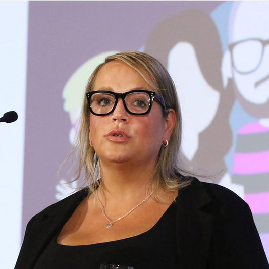 Woman wearing glasses and a black outfit delivering a keynote speech in front of a colourful background