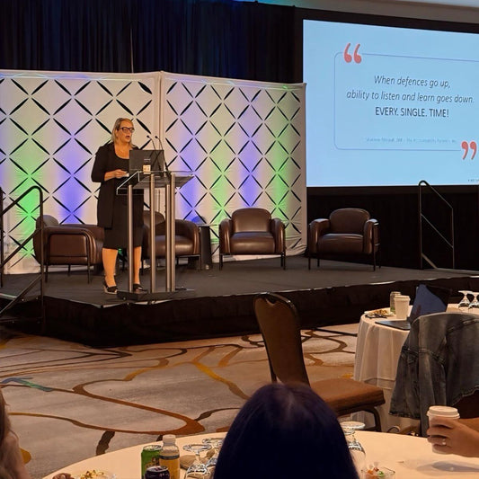 Woman speaking at a podium on a stage with a quote displayed on a screen to the right of her that reads "When defences go up, ability to listen and learn goes down. EVERY. SINGLE. TIME!" - Shannon Rheault