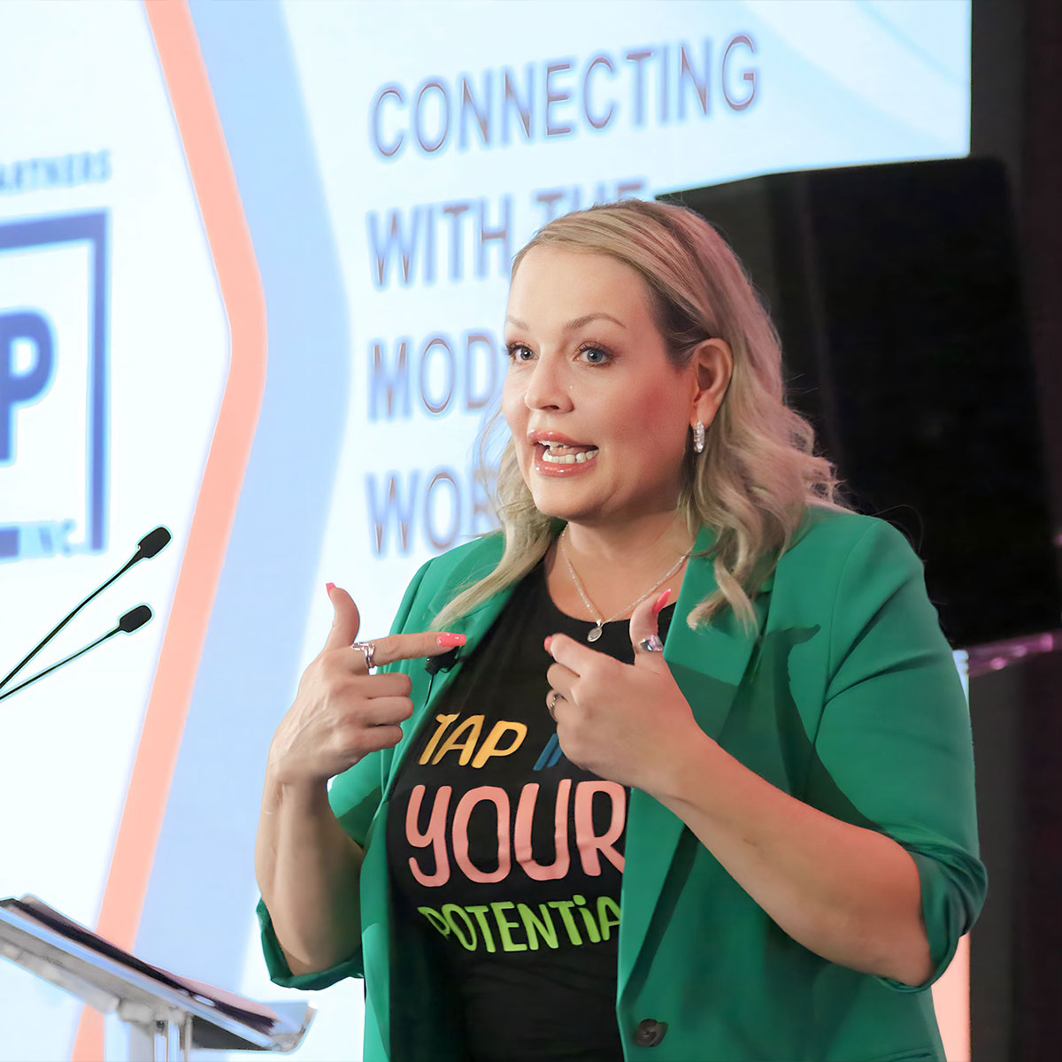 A woman delivering a speech on a stage with a projector behind her, wearing a shirt that reads "Tap into your Potential" with a bright green blazer