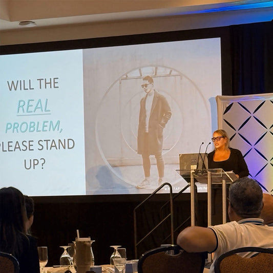 Woman giving a keynote speech and presentation with a screen displaying an image of a person standing with text that reads "will the real problem, please stand up?"