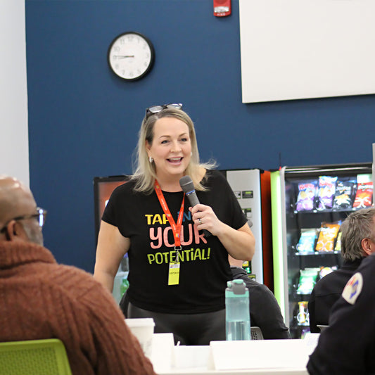 Woman wearing a t shirt that reads "Tap into your potential" teaching and speaking into a microphone in a business classroom with participants at tables around her.