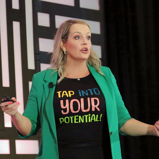 A close up photo of a woman on stage delivering a speech with an animated look on her face. she is wearing a bright green blazer with a black t shirt underneath that reads "Tap into your Potential"