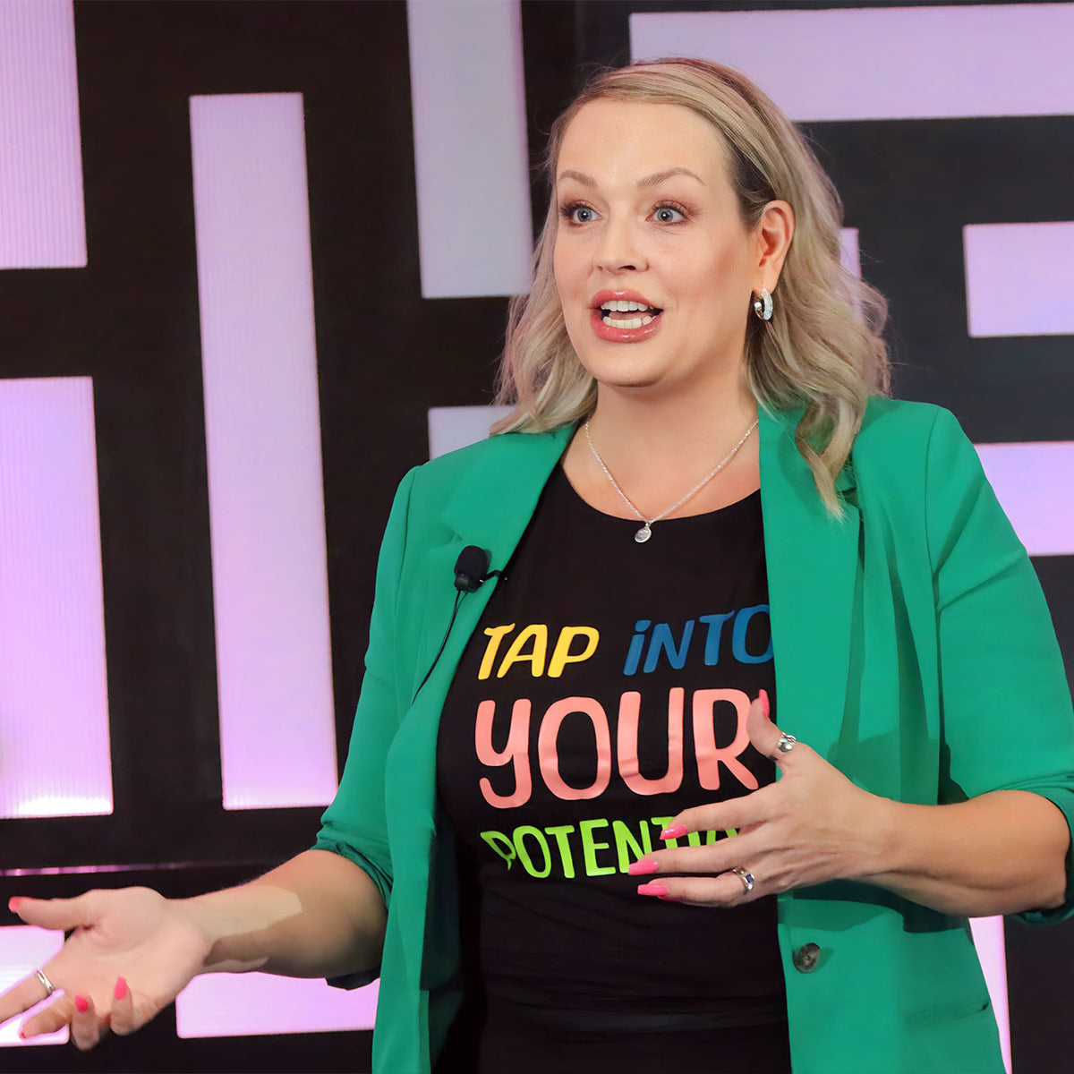 a close up photo of a woman on stage delivering a speech, she is wearing a bright green blazer with a black t shirt underneath that reads "Tap into your Potential"