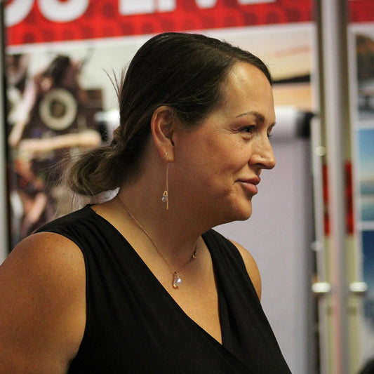 A close up portrait photo of a woman of her left side while she is smiling and looking off to the side. Her hair is in a low bun, she is wearing gold sparkly earrings and necklace with a black top