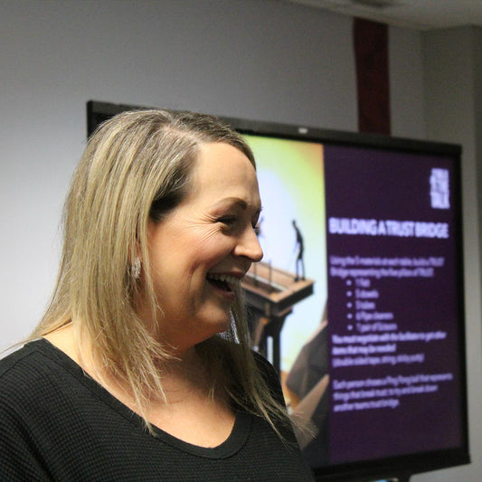 A close up photo of a blonde woman smiling and looking off to the side with a smart board behind her that reads "Building a Trust Bridge"