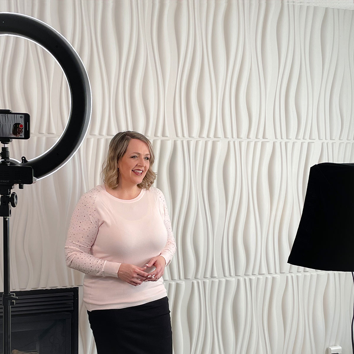 A woman wearing a pink top and black pants standing in front of a white wavy textured wall lit by a ring light. She is looking into a camera and speaking