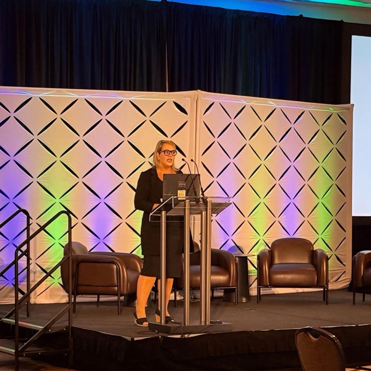 Woman standing at a podium while delivering a keynote speech on a stage with decorative wall and chairs.