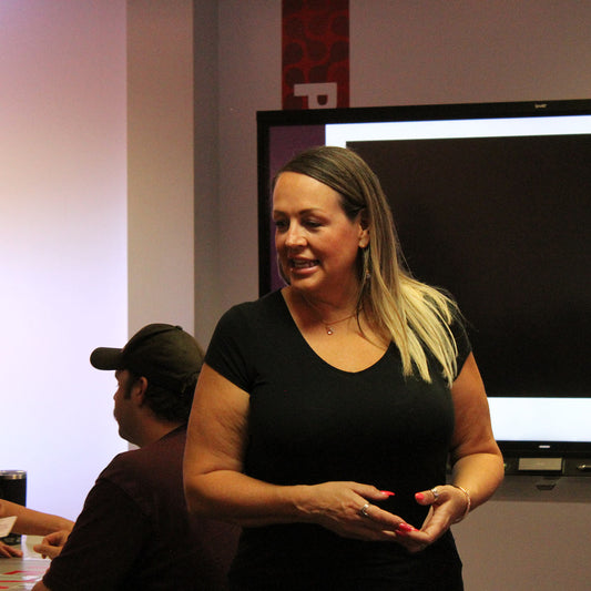 Woman in a black shirt standing and speaking in front of a screen with other people in the background.