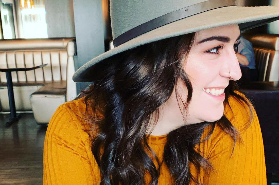close-up photo of a woman wearing an orange top and a grey hat looking off to the side and smiling