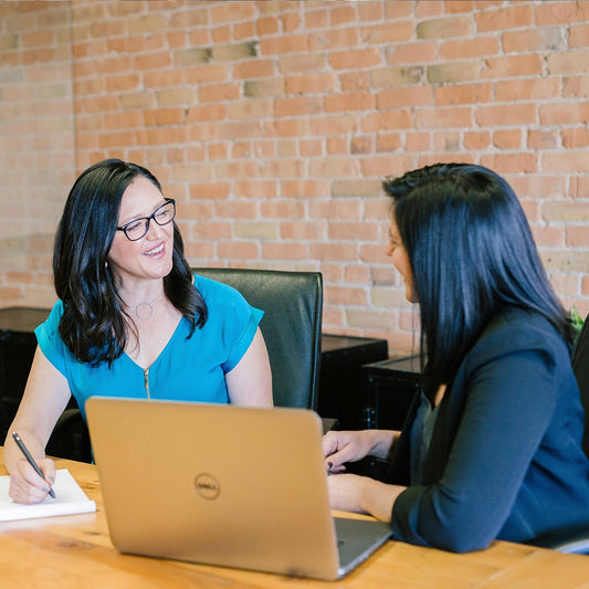 Two women sitting at a table with a laptop, engaged in a conversation, against a brick wall.