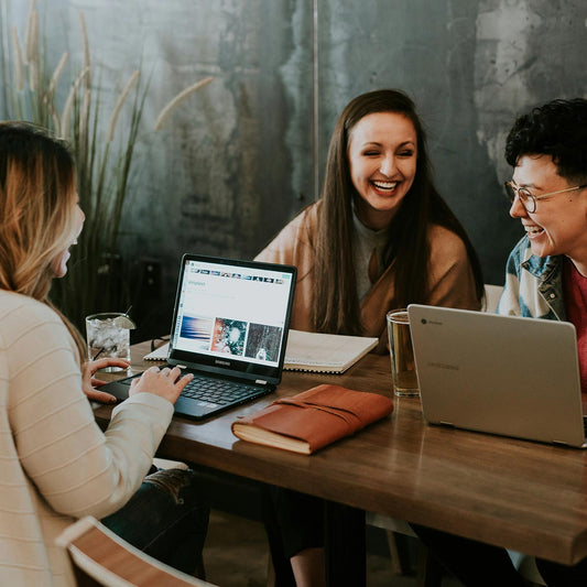 Three people sitting at a table with laptops and a notebook, smiling and engaged in conversation.