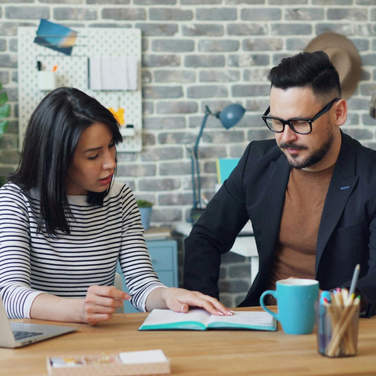 Two people working together at a desk with a brick wall background