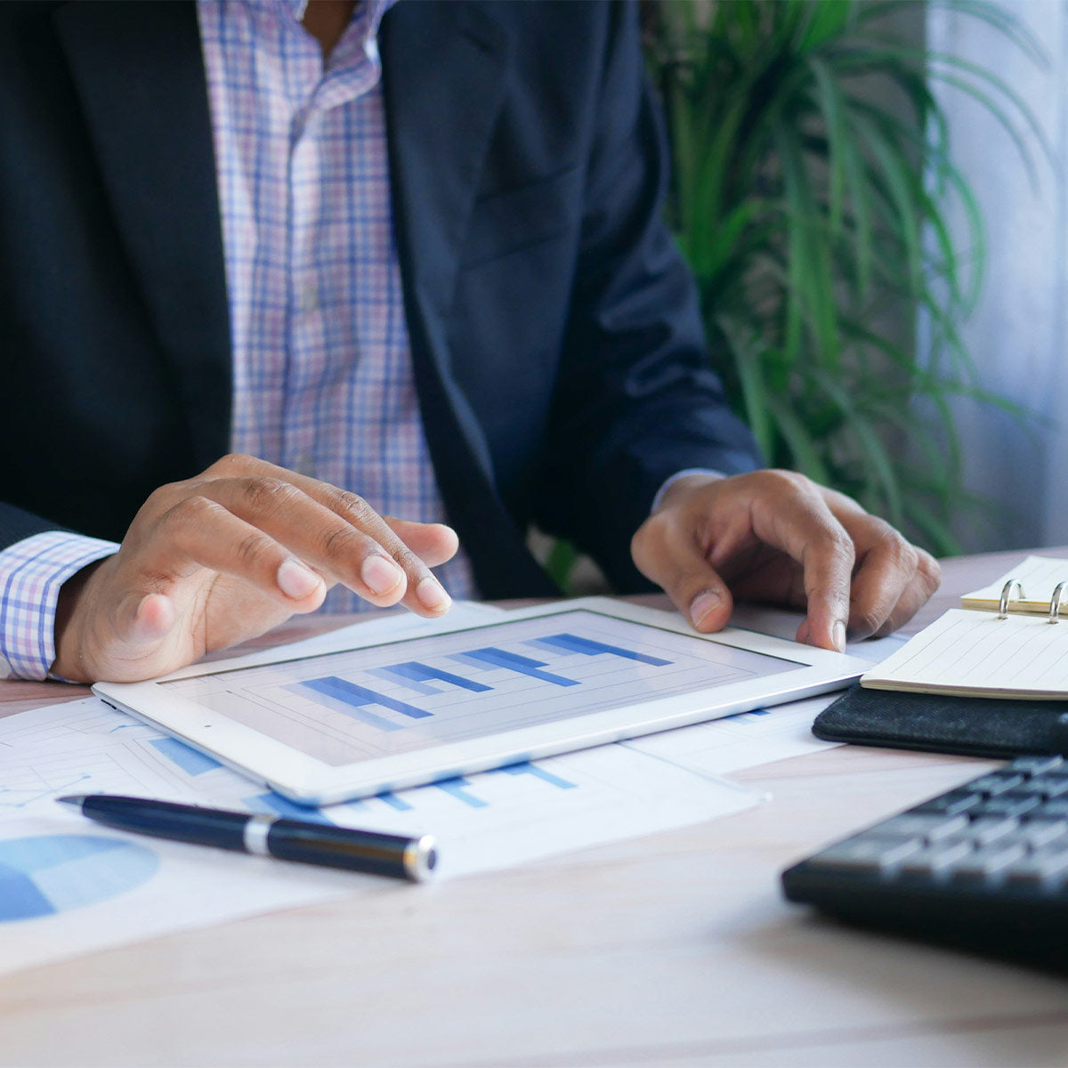 Person using a tablet with financial charts on a desk, with a plant in the background