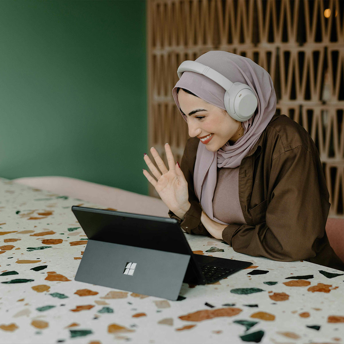 Woman wearing headphones and using a laptop for a video chat on a patterned surface with a green wall and wooden panel in the background.