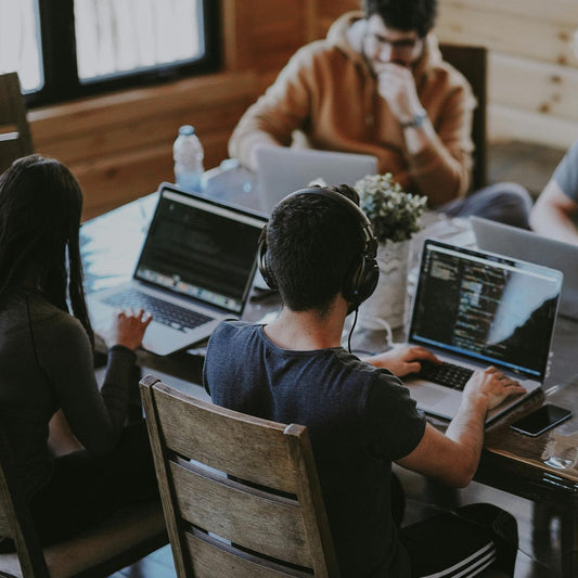 People working on laptops at a table with headphones on, in a casual office setting.