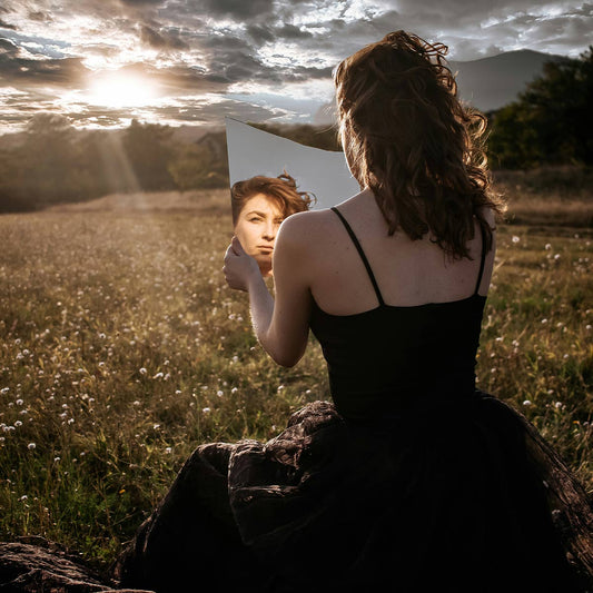 Woman in a black dress holding and looking into a mirror in a field with mountains and sunlit clouds.