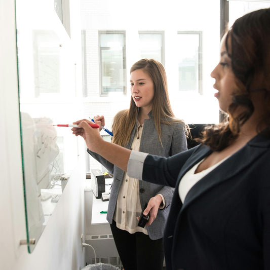 Two women in a professional setting, one writing on a whiteboard with markers.