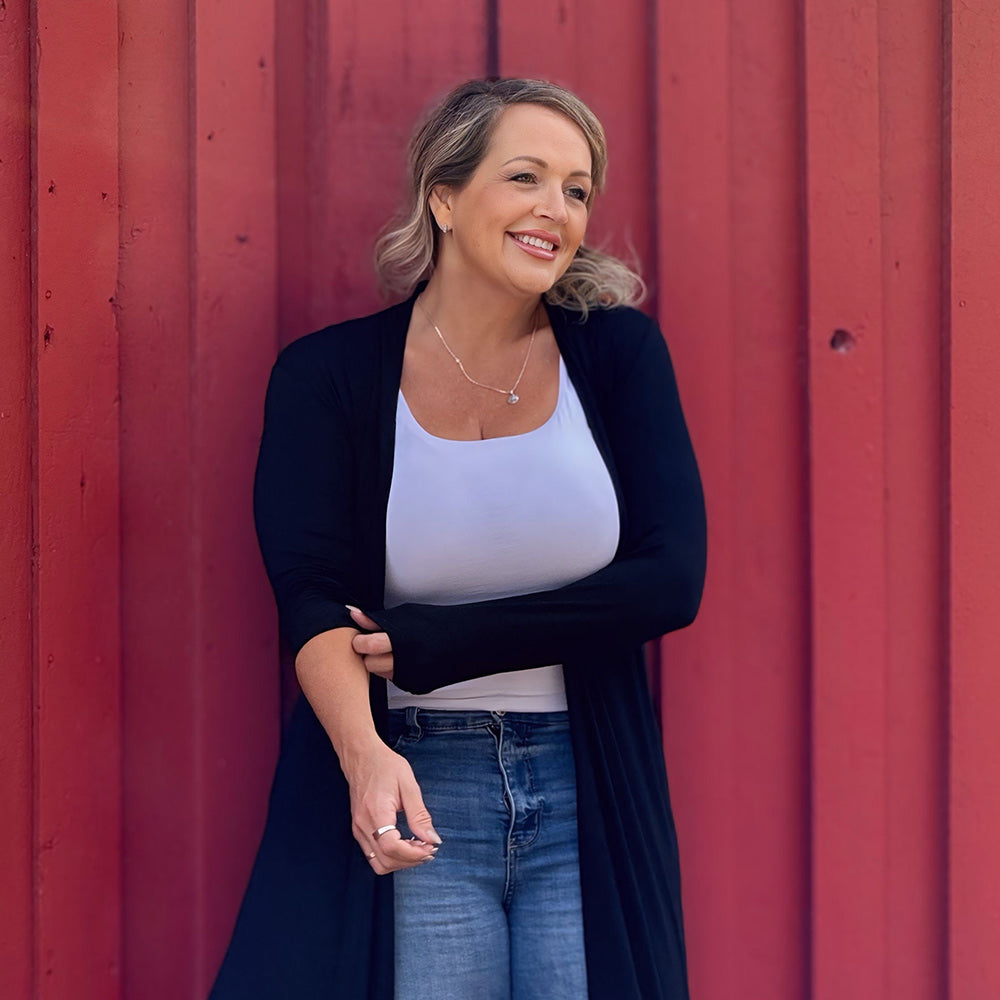 Woman standing against a red wall wearing a black cardigan, white top, and blue jeans.