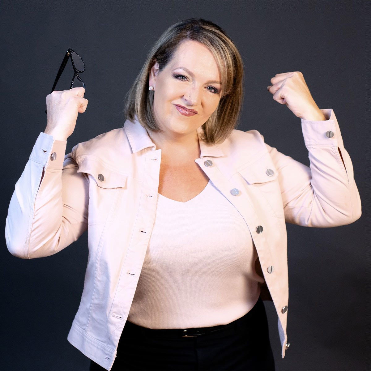 A woman with shoulder length hair wearing a pink shirt and jacket, and black pants; she is holding her fists in the air while holding her glasses and pursing her lips as if she has accomplished a goal