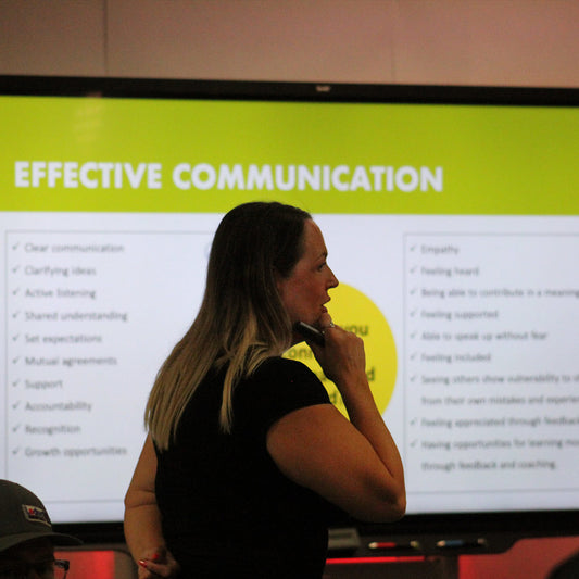 A woman standing with her hand on her chin while mid sentence, in front of a smart board that reads "Effective Communication" as a title with rows of examples below it. 