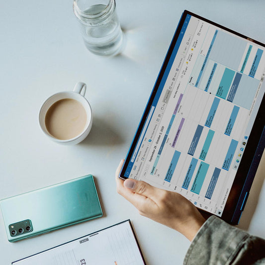 Person holding a tablet with a productivity app on a desk with a phone, coffee, and water bottle.