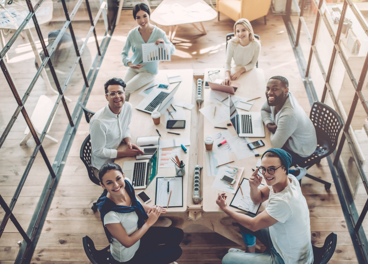 group of people in business setting sitting at a long table with laptops, papers, cell phones, writing utensils, etc. smiling and looking up to the camera angle 
